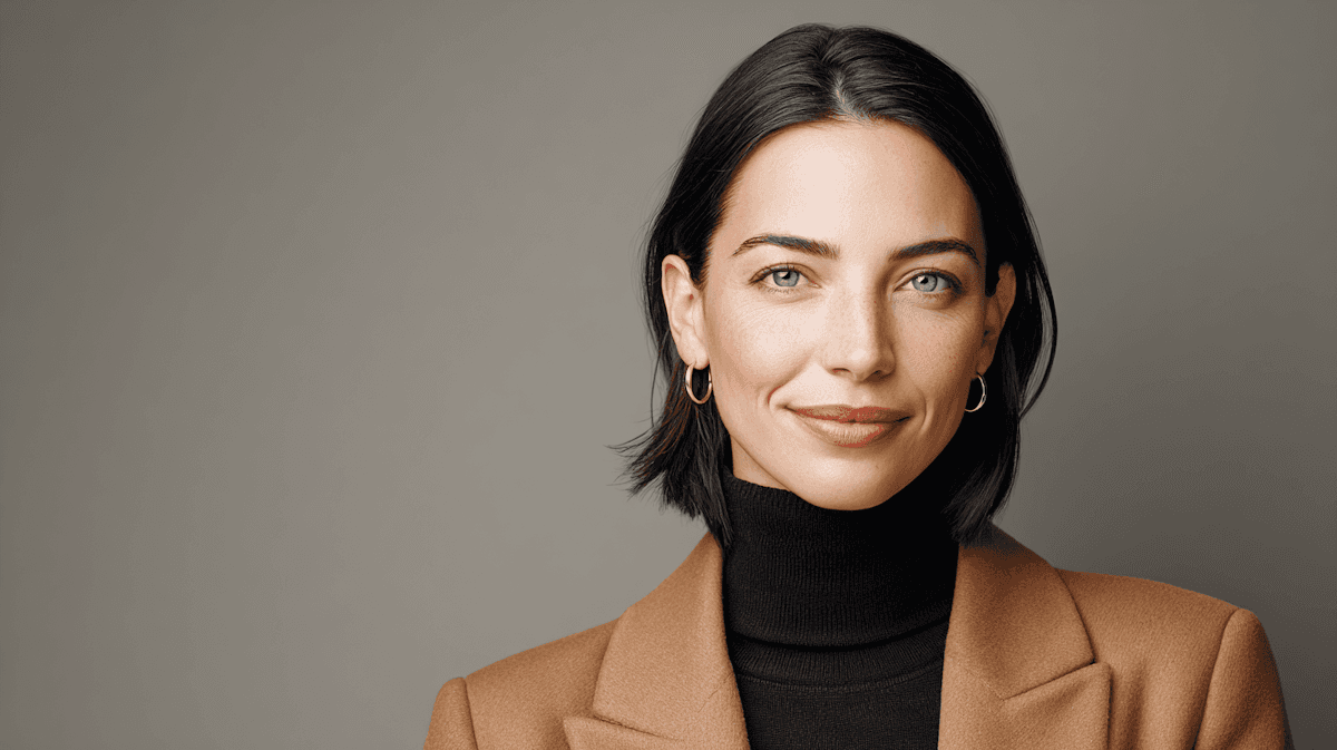 Professional headshot of a woman with short dark hair wearing a camel blazer and black turtleneck against a gray background.